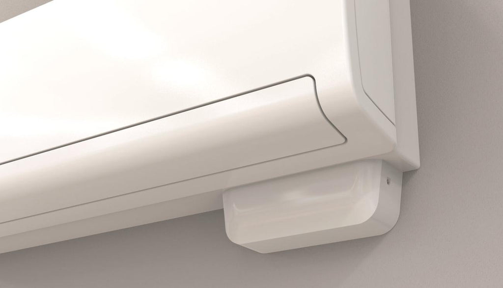 A close-up of a white wall-mounted air conditioner or split system indoor unit with a small white receiver/dryer box below it against a light gray wall.