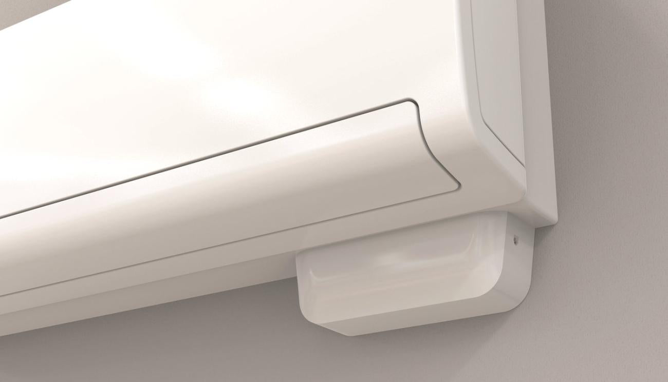 A close-up of a white wall-mounted air conditioner or split system indoor unit with a small white receiver/dryer box below it against a light gray wall.